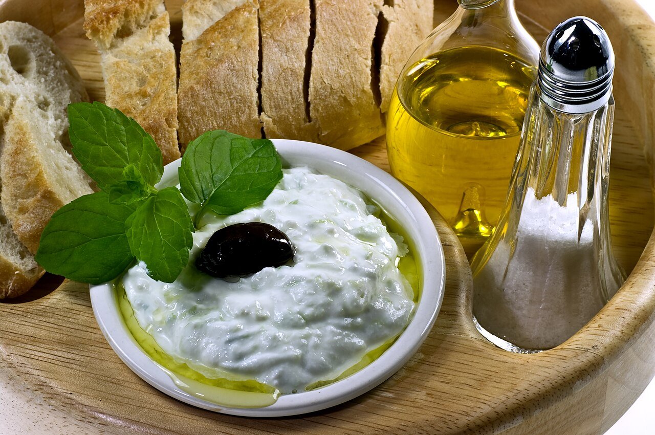 Photograph of cucumber yogurt dip with mint and a black olive, served beside bread