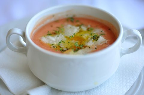 Red gazpacho in a white handled bowl with oil and pale garnish